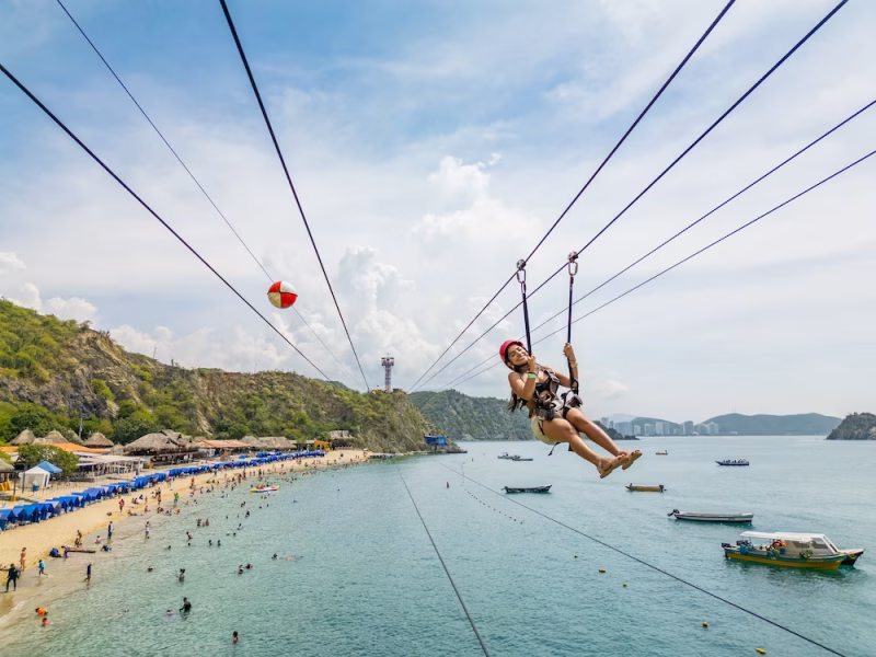 Turista deslizándose en canopy sobre la bahía de Playa Blanca en Cartagena, con la playa llena de visitantes y el mar turquesa de fondo